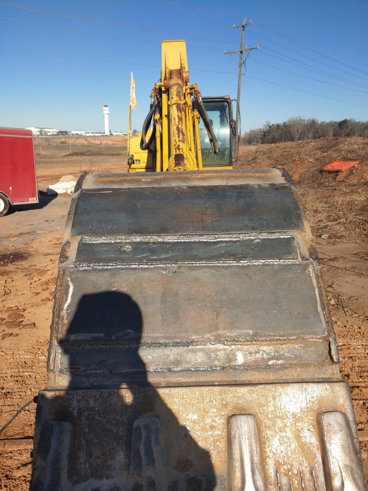 Fresh weld beads laid across the back of an excavator bucket at a red-dirt jobsite, the excavator parked behind it.