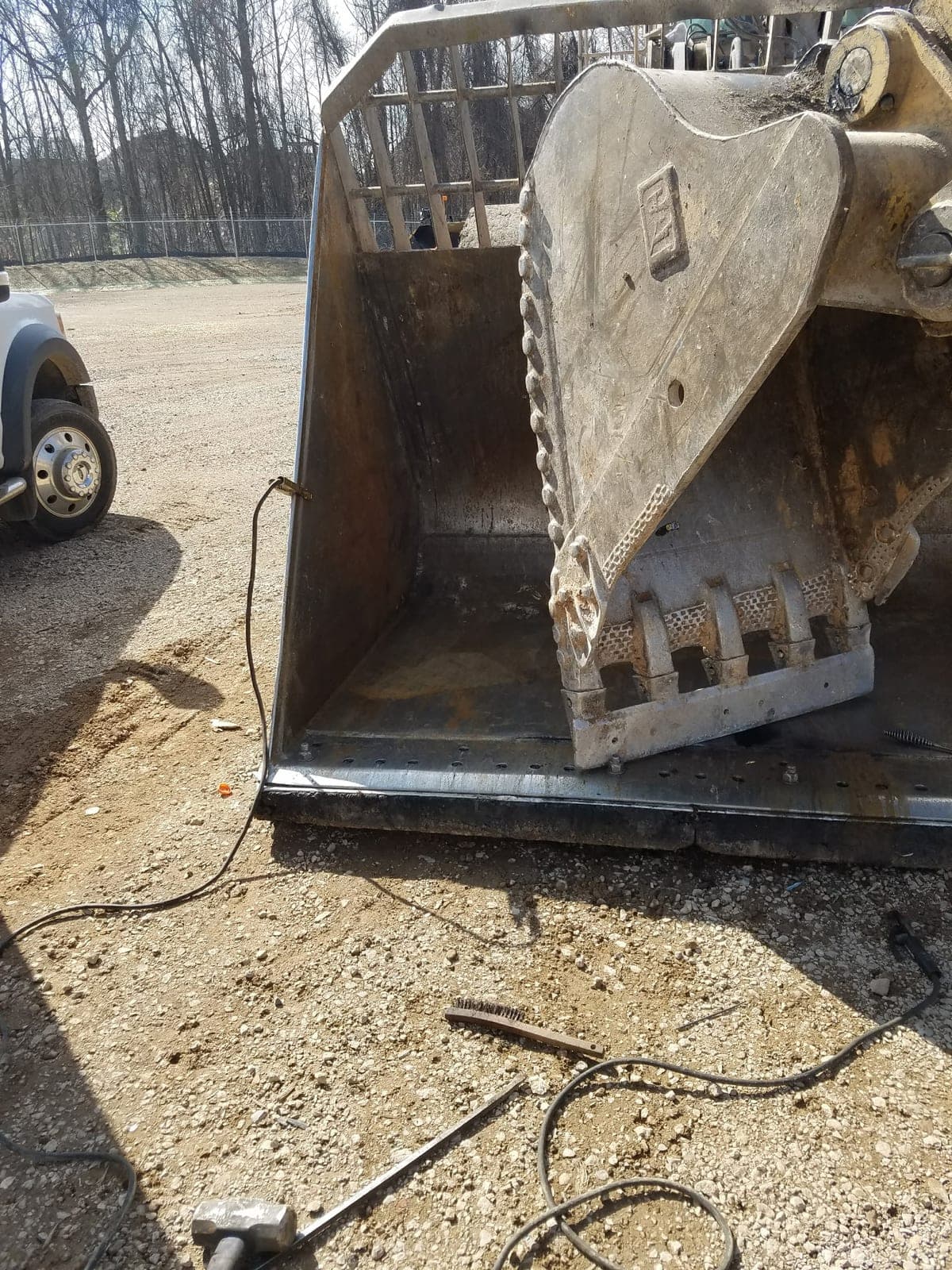 A Caterpillar bucket in the gravel yard with a replacement cutting edge being fitted, welding lead and sledgehammer on the ground.