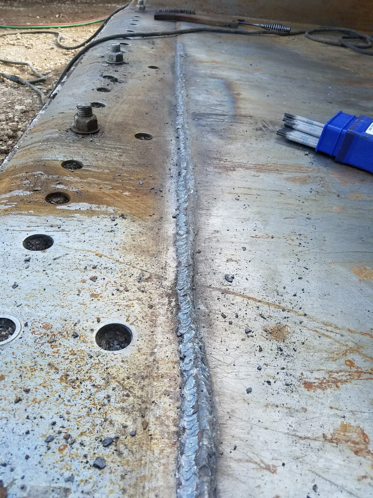 A long weld bead run down the length of a bucket cutting edge, with a box of 7018 rod laid out alongside.