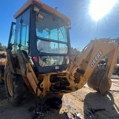 A John Deere backhoe loader with the side panels opened up, parked in the yard under bright sun.