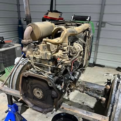 A diesel engine removed from its chassis and resting on a frame stand in the shop, exhaust manifold and turbo still attached.