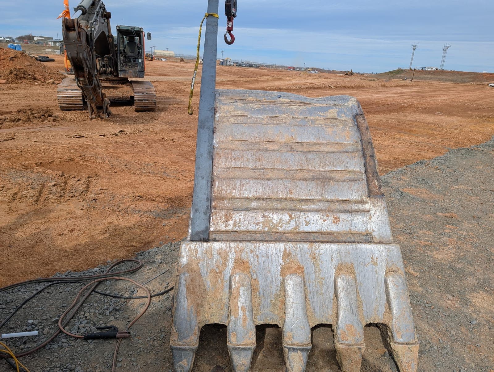 The rebuilt excavator bucket slung upright on a jobsite, welding leads coiled on the ground and the excavator parked behind.