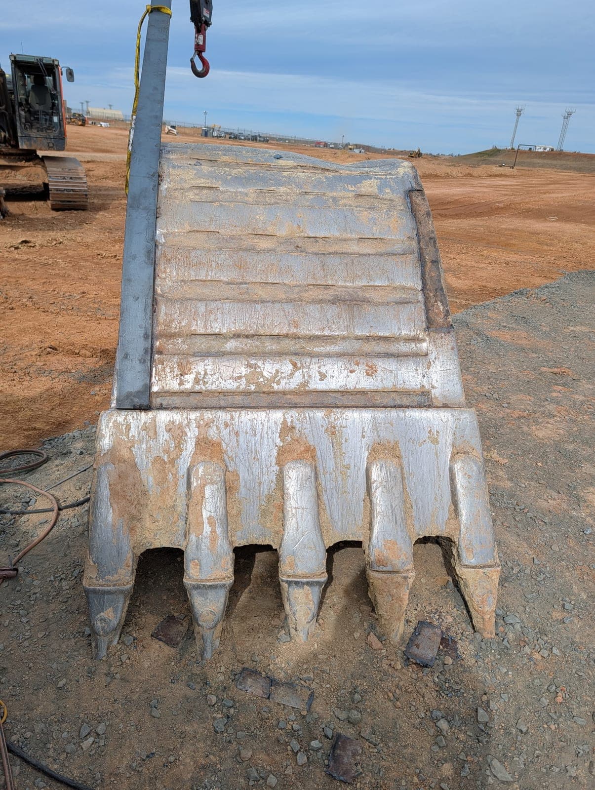 An excavator bucket standing upright on the gravel with rebuilt wear surfaces and teeth, slung from the crane hook.