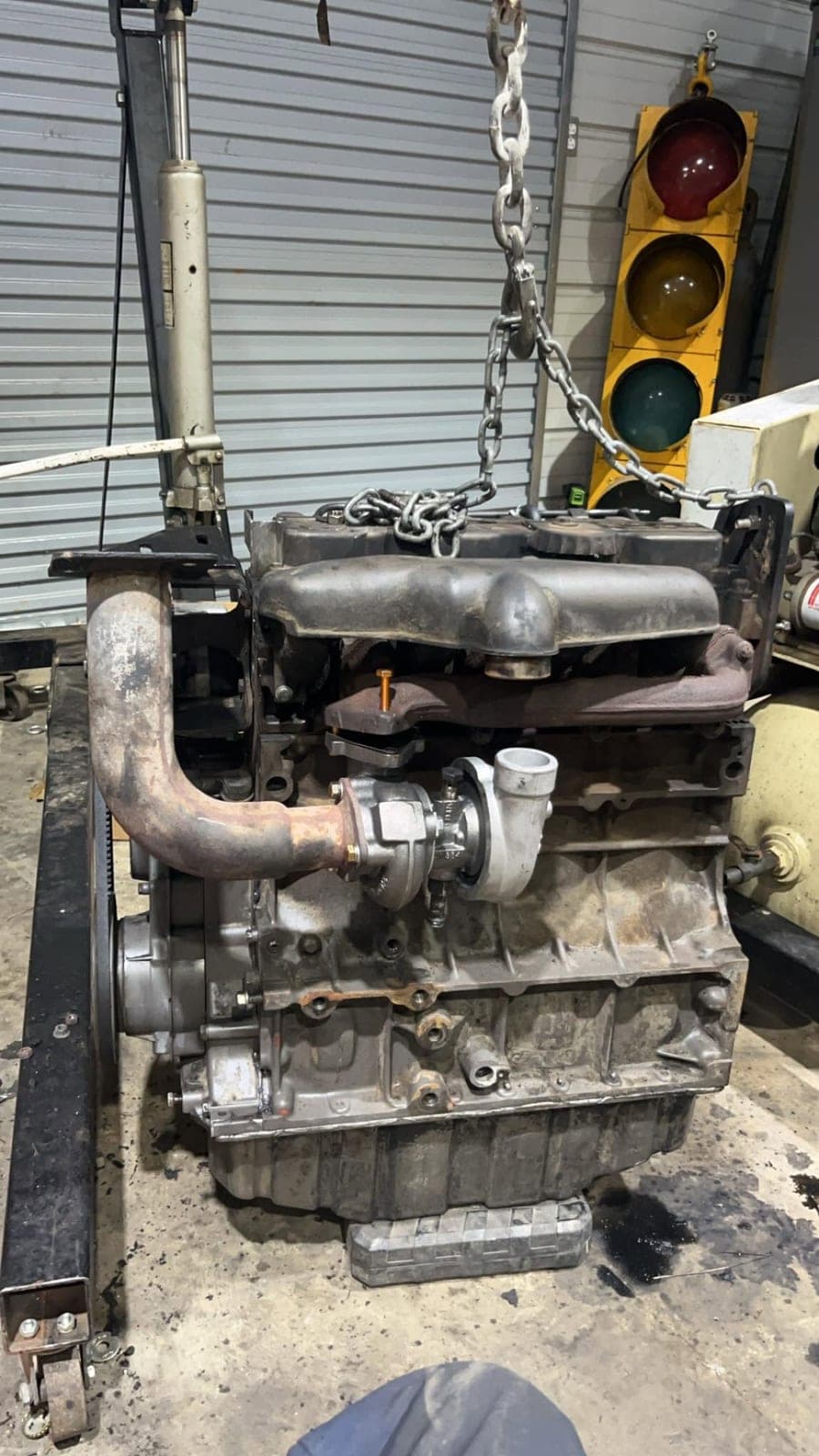 A rebuilt four-cylinder diesel engine with turbocharger and exhaust elbow hanging on a chain hoist outside the shop bay.