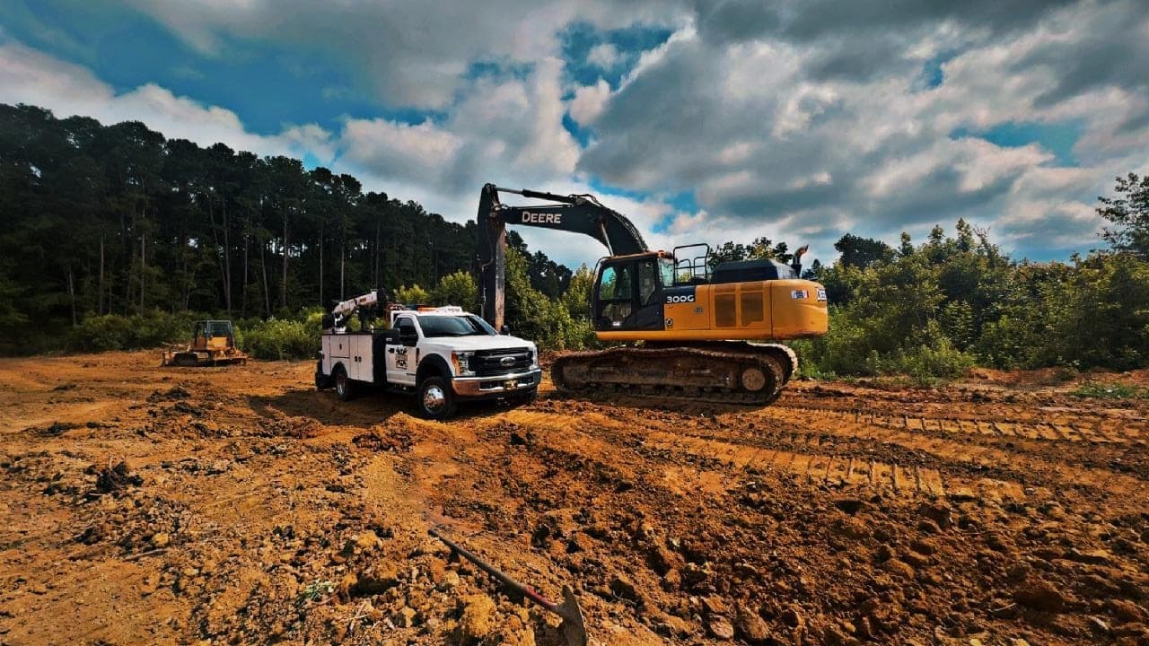 The Joyner Services service truck parked beside a John Deere 300G excavator on a red-dirt jobsite under a big cloudy sky.