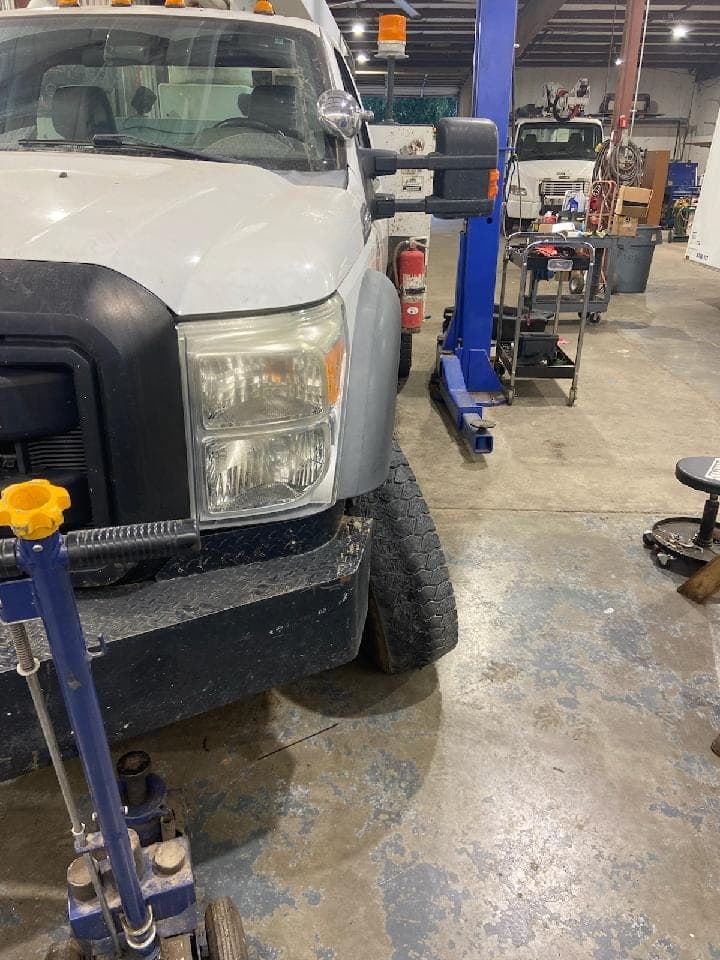 The front of the white Ford service truck up on a floor jack inside the shop bay, with a transmission jack alongside.