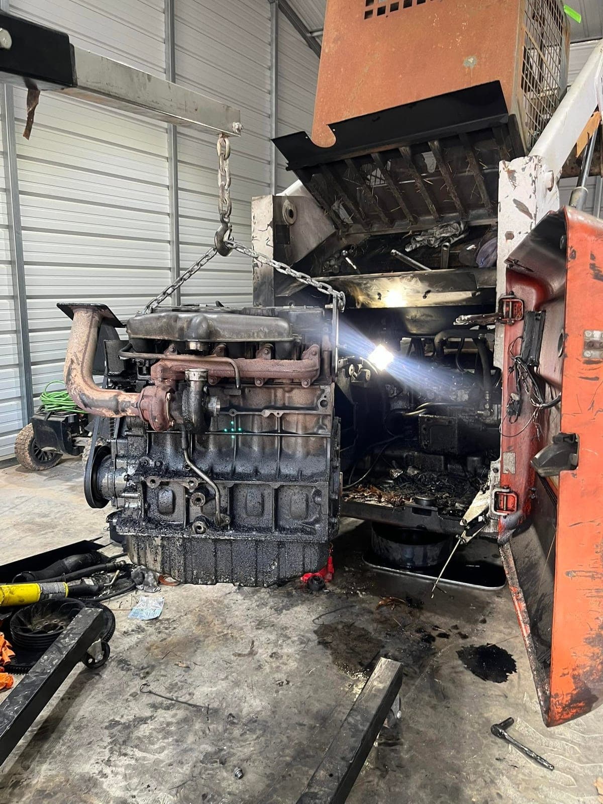 A diesel engine lifted out of an orange skid steer loader on a chain hoist, sitting on the shop floor beside the stripped machine.