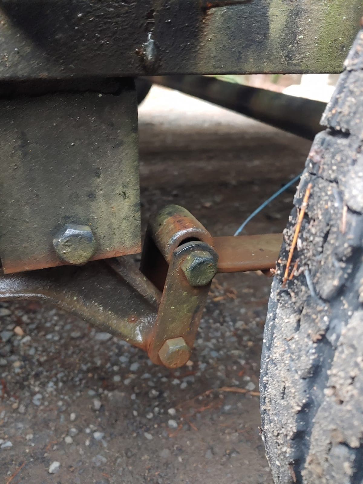 A rusted spring shackle and hanger bracket under a machine, beside a mud-packed tire.