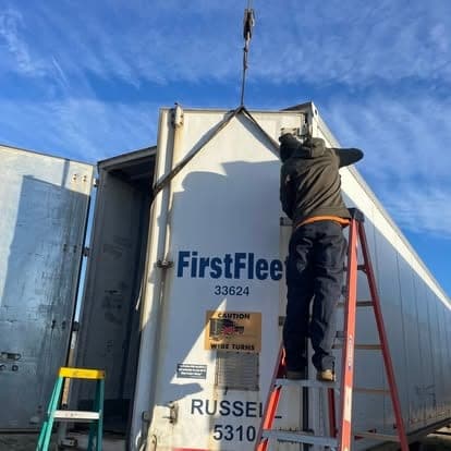 A technician on a ladder repairing the upper side panel of a white FirstFleet dry van trailer.