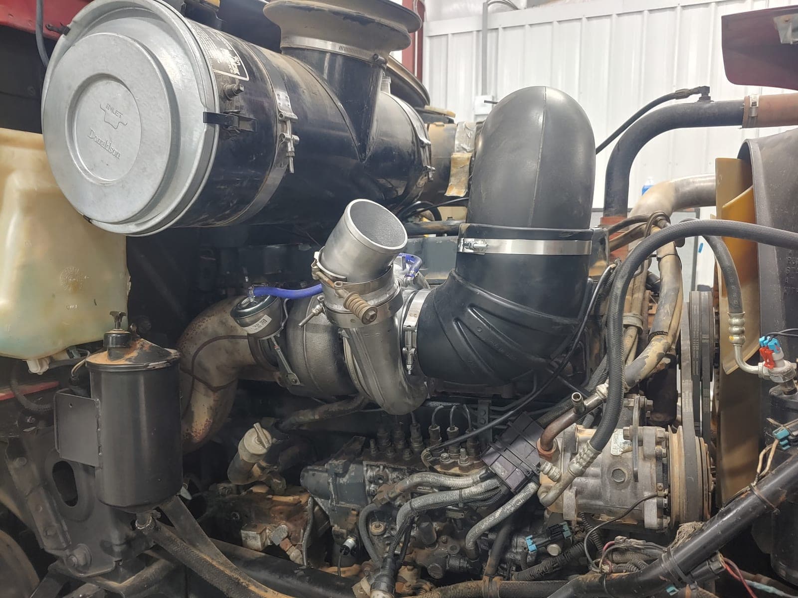 A turbocharger and new intake elbow clamped up in the engine bay of a truck in the shop, air cleaner and belt drive alongside.