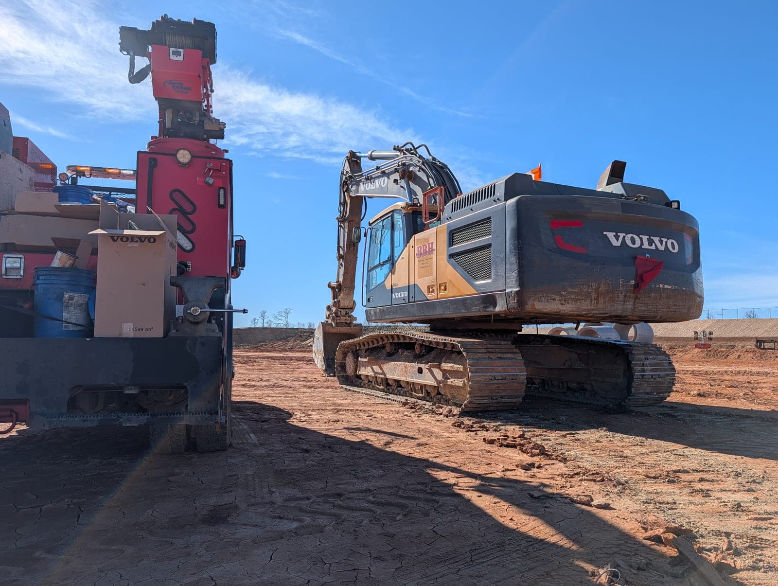 A Volvo tracked excavator on a red-dirt jobsite with the red mechanic service truck and its crane parked alongside.